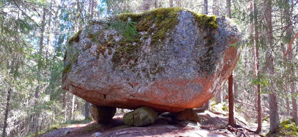 Metsässä on iso siirtolohkare kallion päällä. Sammaleinen iso kivi lepää pienempien kivien päällä, mutta pysyy kuitenkin paikallaan,